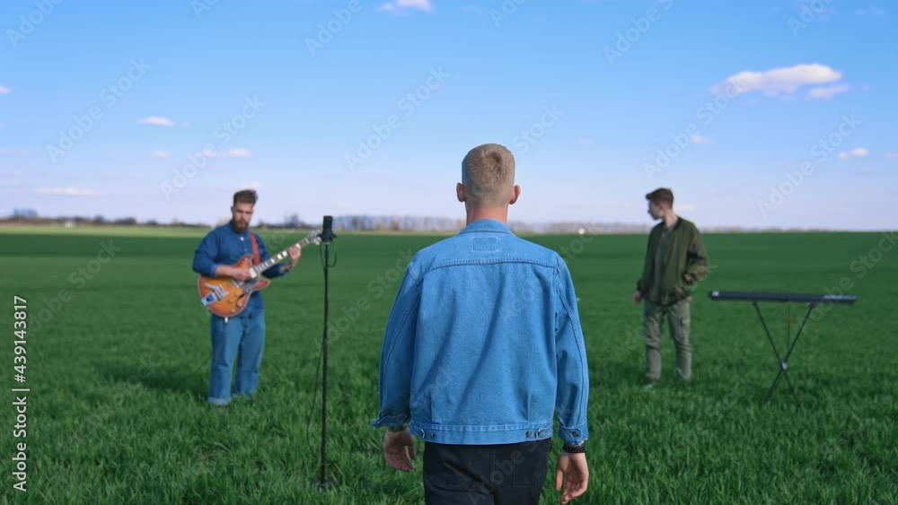 Music band on green field background. Young men playing musical instruments outdoors. Man singer coming to a microphone. Music band on green field background. Young men playing musical instruments outdoors. Man singer coming to a microphone.