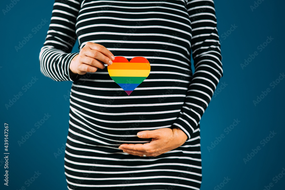 Closeup of pregnant woman holding a rainbow colored hearth against a black and white striped dress and blue studio background image