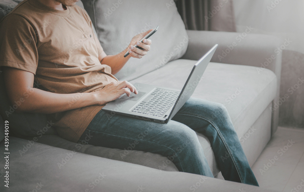 Fototapeta premium young man typing on mobile phone while sitting on a sofa at home with laptop computer. Man using his smartphone for online banking.work from home concept.