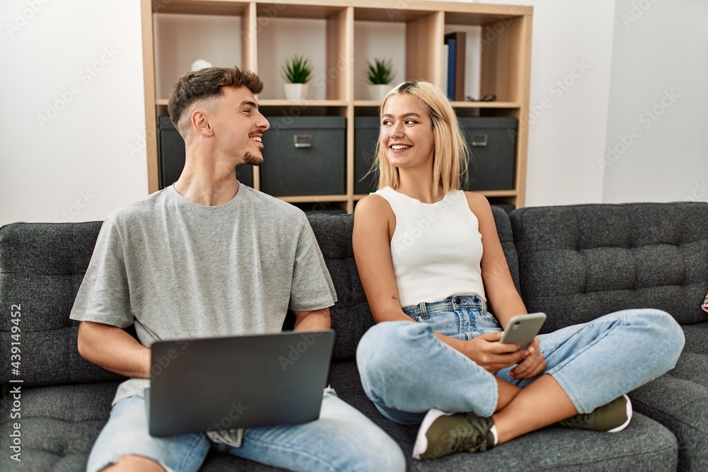 Young caucasian couple smiling happy using laptop and smartphone sitting on the sofa at home. Young caucasian couple smiling happy using laptop and smartphone sitting on the sofa at home.