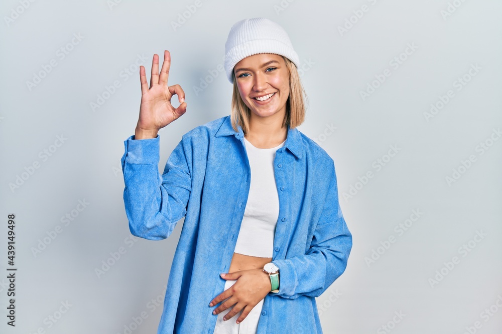 Beautiful blonde woman wearing wool hat smiling positive doing ok sign with hand and fingers. successful expression. Beautiful blonde woman wearing wool hat smiling positive doing ok sign with hand and fingers. successful expression.