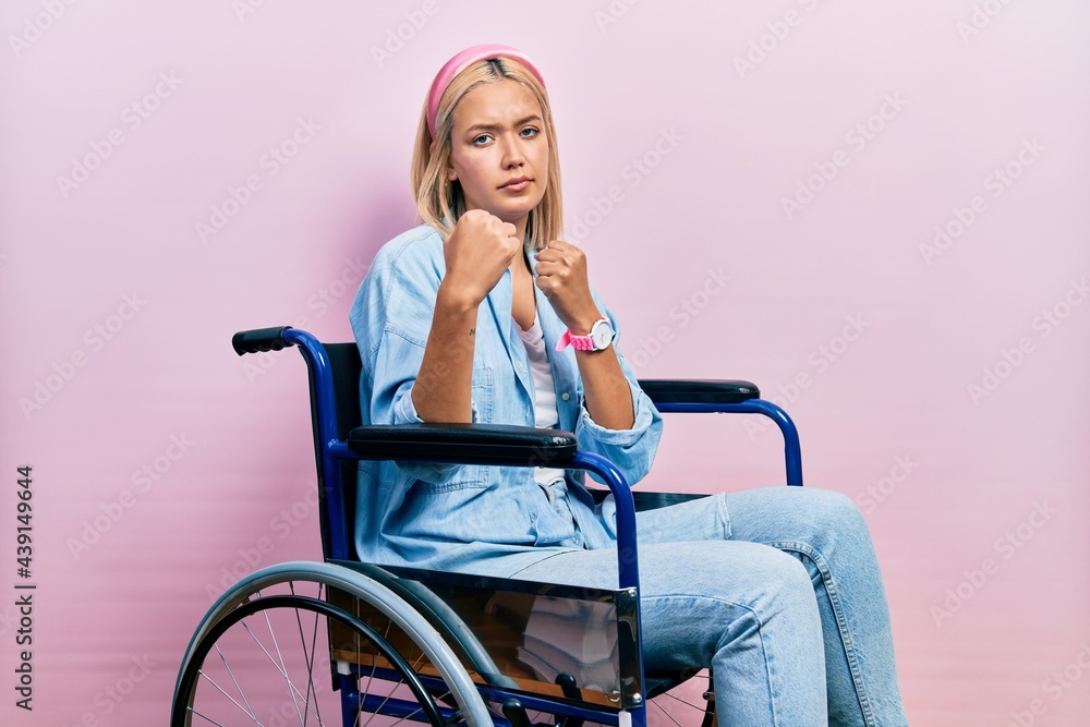 Beautiful blonde woman sitting on wheelchair ready to fight with fist defense gesture, angry and upset face, afraid of problem Beautiful blonde woman sitting on wheelchair ready to fight with fist defense gesture, angry and upset face, afraid of problem