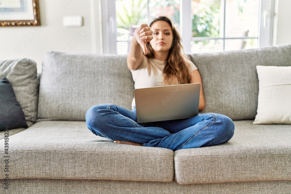 Beautiful young brunette woman sitting on the sofa using computer laptop at home looking unhappy and angry showing rejection and negative with thumbs down gesture. bad expression. Beautiful young brunette woman sitting on the sofa using computer laptop at home looking unhappy and angry showing rejection and negative with thumbs down gesture. bad expression.