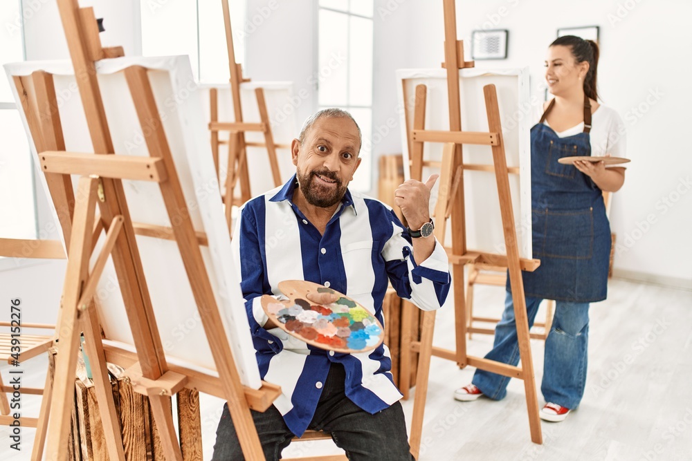 Senior artist man at art studio smiling with happy face looking and pointing to the side with thumb up. Senior artist man at art studio smiling with happy face looking and pointing to the side with thumb up.