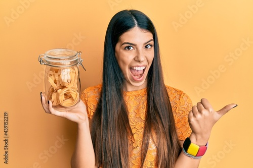 Young hispanic woman holding jar with uncooked pasta pointing thumb up to the side smiling happy with open mouth