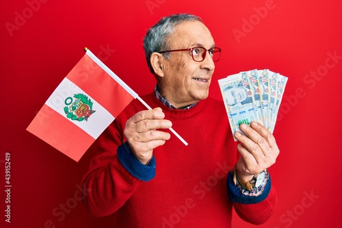 Handsome senior man with grey hair holding peru flag and peruvian sol banknotes smiling looking to the side and staring away thinking.