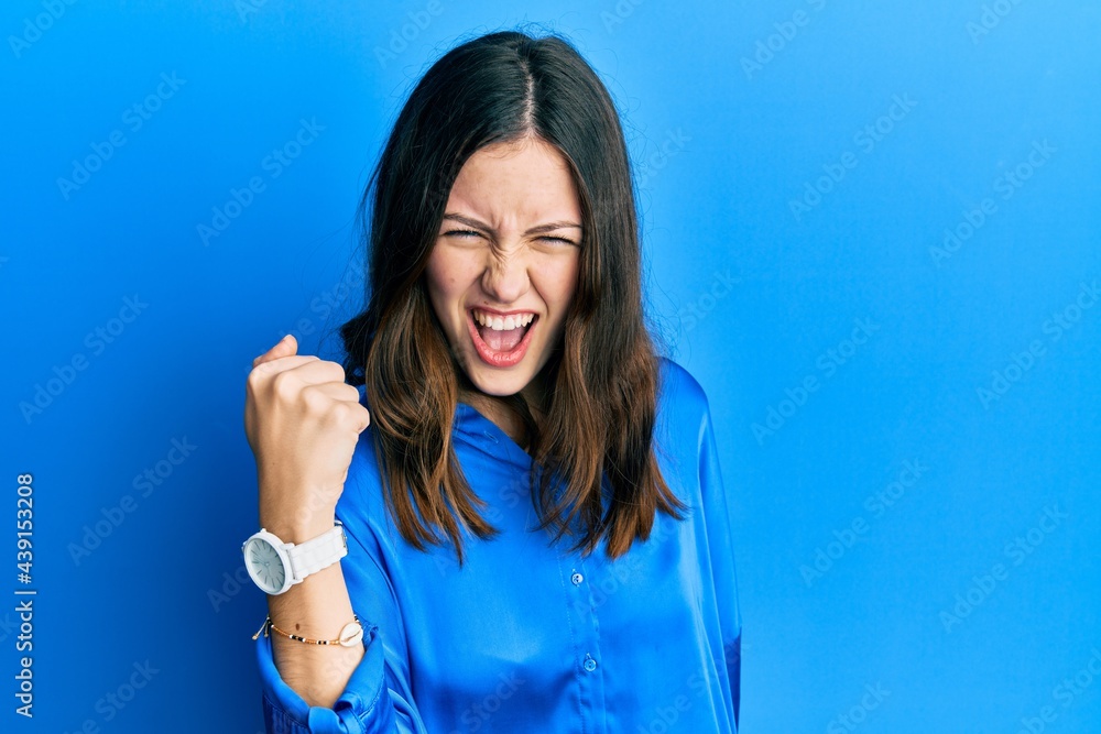 Fototapeta premium Young brunette woman wearing casual blue shirt angry and mad raising fist frustrated and furious while shouting with anger. rage and aggressive concept.