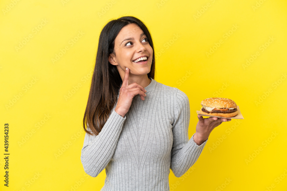 Young caucasian woman holding a burger isolated on yellow background thinking an idea while looking up Young caucasian woman holding a burger isolated on yellow background thinking an idea while looking up