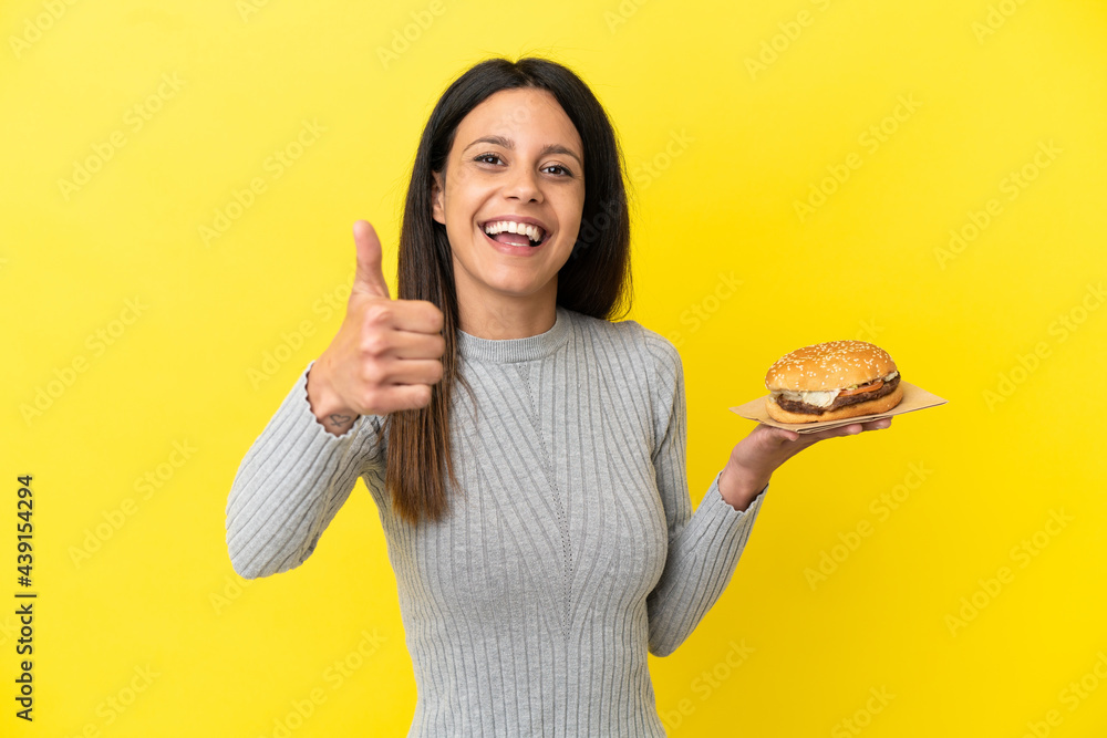 Young caucasian woman holding a burger isolated on yellow background with thumbs up because something good has happened Young caucasian woman holding a burger isolated on yellow background with thumbs up because something good has happened