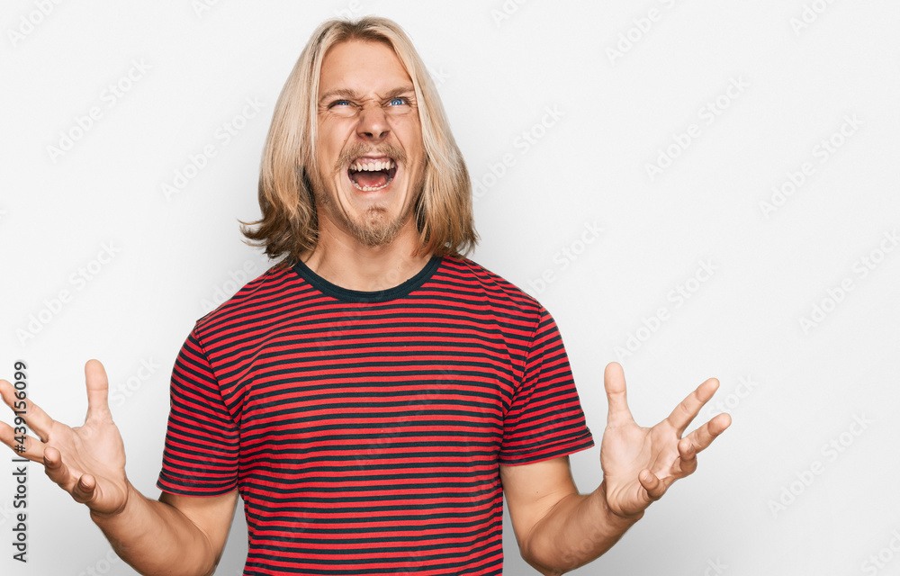 Caucasian man with blond long hair wearing casual striped t shirt crazy and mad shouting and yelling with aggressive expression and arms raised. frustration concept. Caucasian man with blond long hair wearing casual striped t shirt crazy and mad shouting and yelling with aggressive expression and arms raised. frustration concept.