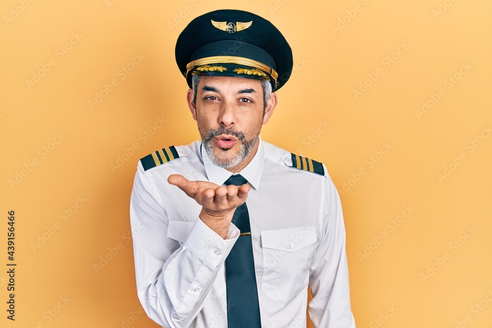 Handsome middle age man with grey hair wearing airplane pilot uniform looking at the camera blowing a kiss with hand on air being lovely and sexy. love expression. Handsome middle age man with grey hair wearing airplane pilot uniform looking at the camera blowing a kiss with hand on air being lovely and sexy. love expression.