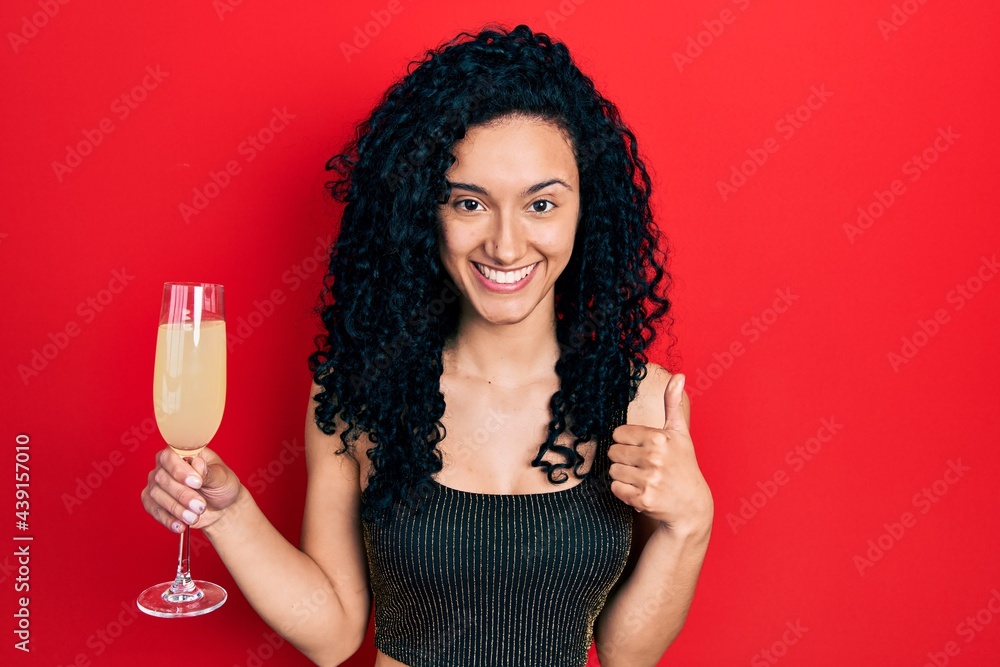 Young hispanic woman with curly hair drinking a glass of sparkling champagne smiling happy and positive, thumb up doing excellent and approval sign Young hispanic woman with curly hair drinking a glass of sparkling champagne smiling happy and positive, thumb up doing excellent and approval sign