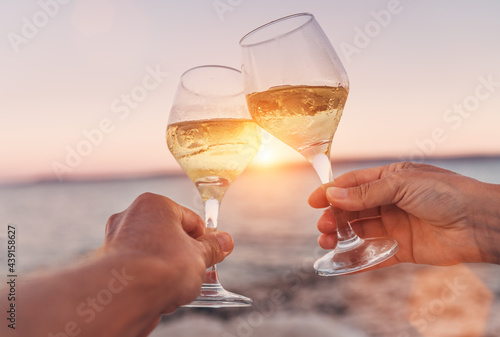Couple in love clinking with white wine glasses while they enjoying the beautiful pink seaside sunset. Romantic vacation or dating concept close-up image.