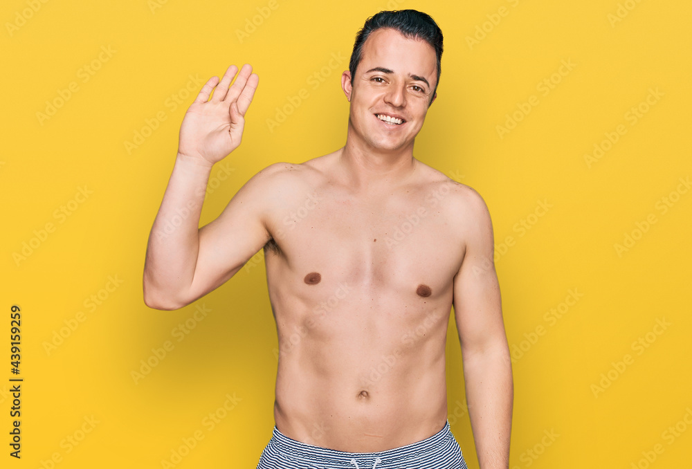 Handsome young man wearing swimwear shirtless waiving saying hello happy and smiling, friendly welcome gesture