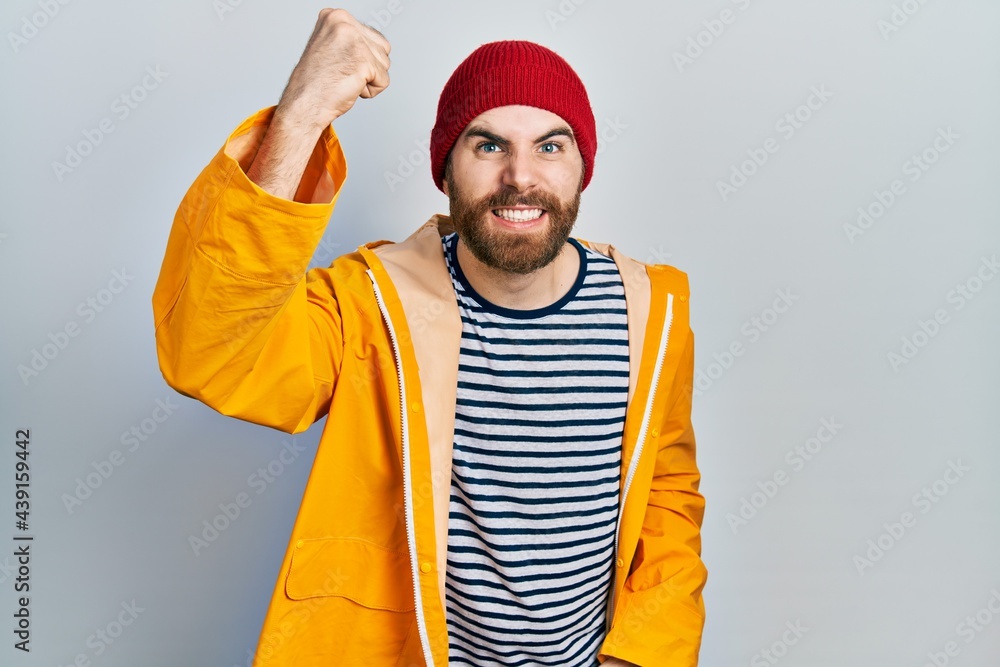 Caucasian man with beard wearing yellow raincoat angry and mad raising fist frustrated and furious while shouting with anger. rage and aggressive concept. Caucasian man with beard wearing yellow raincoat angry and mad raising fist frustrated and furious while shouting with anger. rage and aggressive concept.