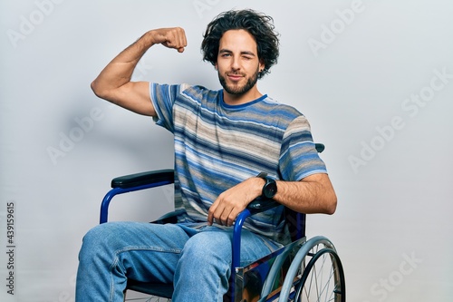 Handsome hispanic man sitting on wheelchair strong person showing arm muscle, confident and proud of power