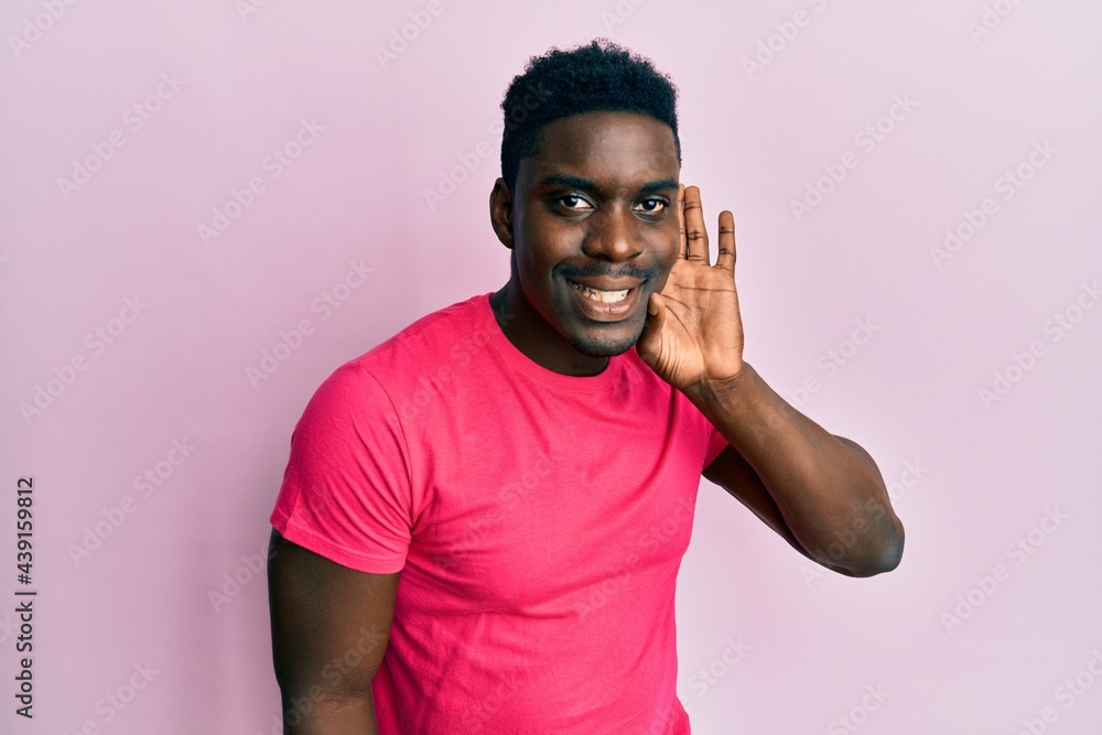 Handsome black man wearing casual pink t shirt smiling with hand over ear listening and hearing to rumor or gossip. deafness concept. Handsome black man wearing casual pink t shirt smiling with hand over ear listening and hearing to rumor or gossip. deafness concept.