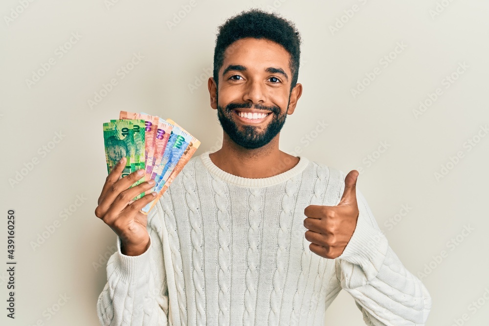 Handsome hispanic man with beard holding south african rand banknotes smiling happy and positive, thumb up doing excellent and approval sign Handsome hispanic man with beard holding south african rand banknotes smiling happy and positive, thumb up doing excellent and approval sign