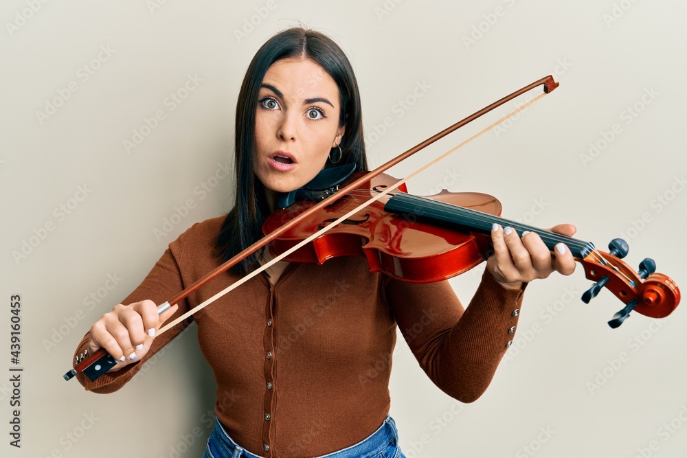 Young brunette woman playing violin afraid and shocked with surprise and amazed expression, fear and excited face. Young brunette woman playing violin afraid and shocked with surprise and amazed expression, fear and excited face.