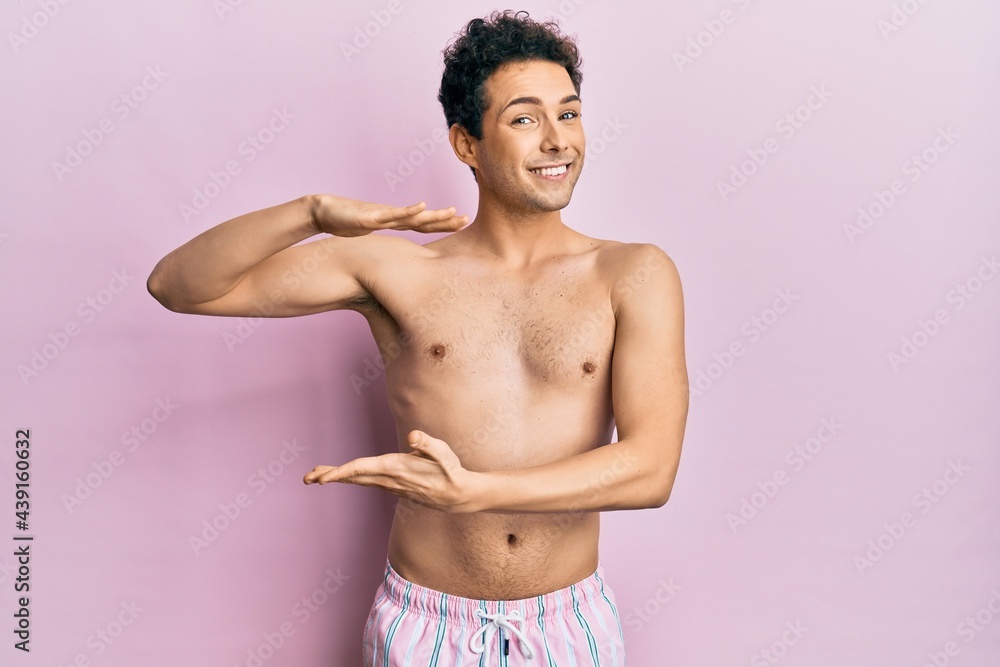 Fototapeta premium Young handsome man wearing swimwear shirtless gesturing with hands showing big and large size sign, measure symbol. smiling looking at the camera. measuring concept.