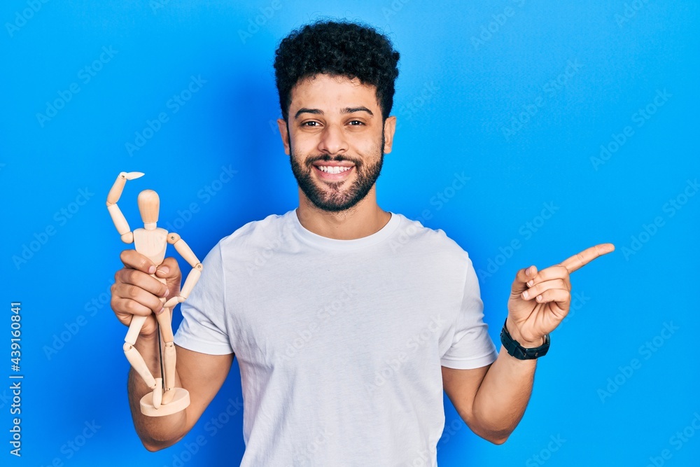 Young arab man with beard holding small wooden manikin smiling happy pointing with hand and finger to the side Young arab man with beard holding small wooden manikin smiling happy pointing with hand and finger to the side