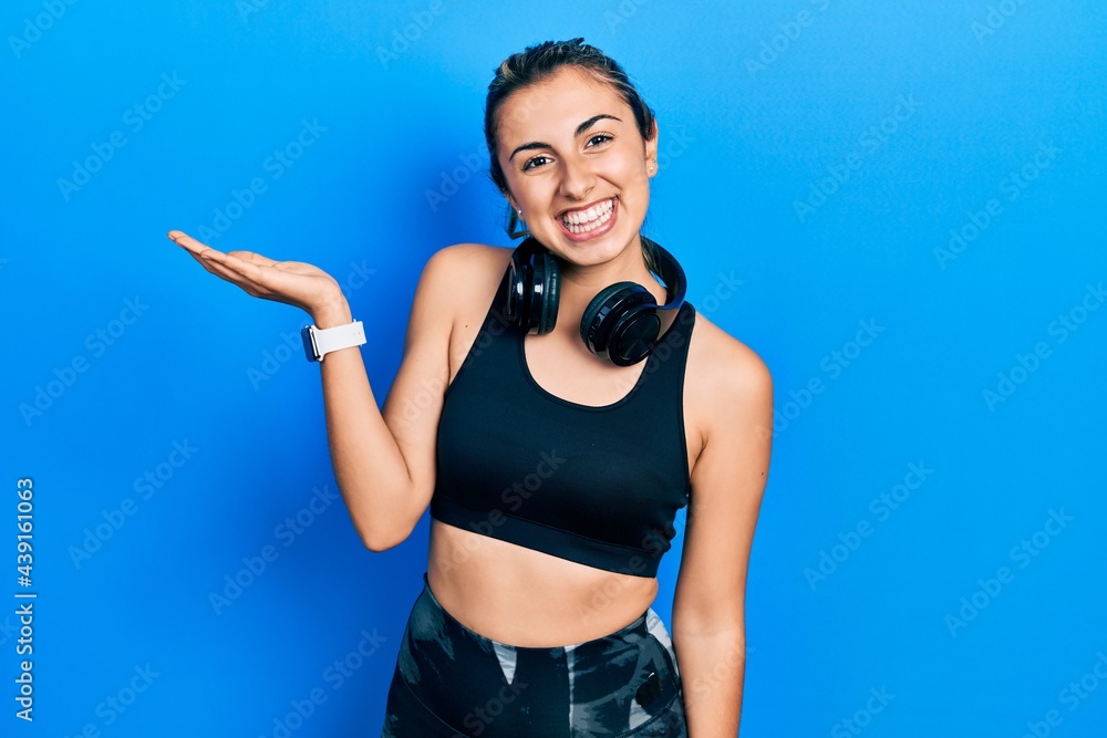 Beautiful hispanic woman wearing gym clothes and using headphones smiling cheerful presenting and pointing with palm of hand looking at the camera.