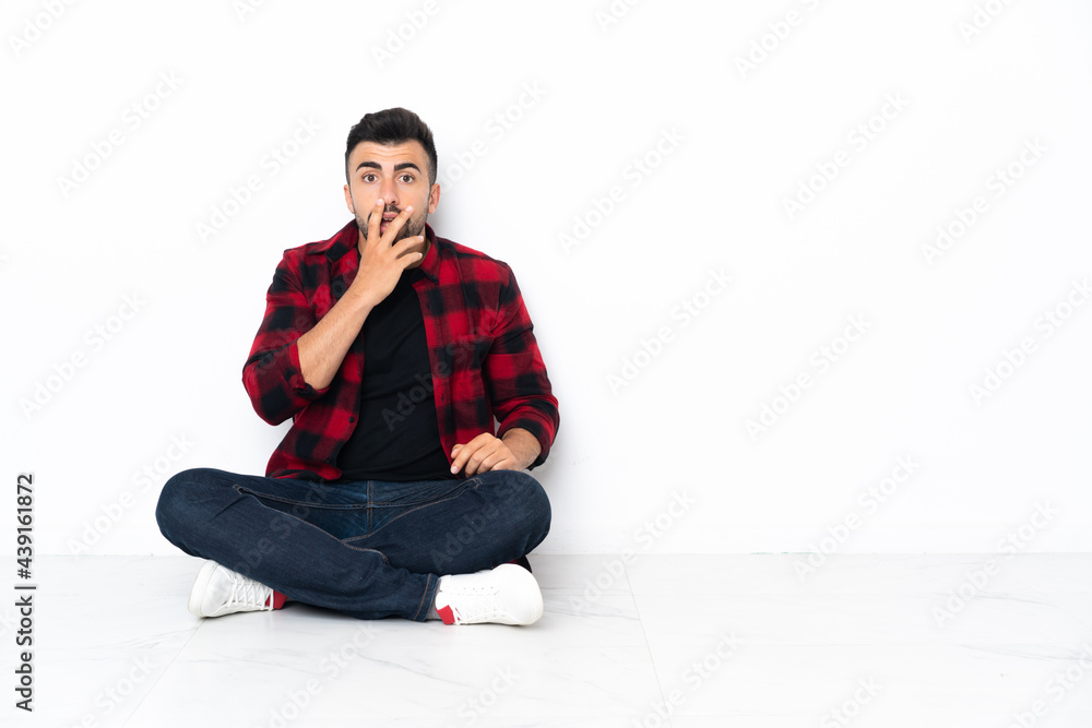 Young handsome man sitting on the floor surprised and shocked while looking right Young handsome man sitting on the floor surprised and shocked while looking right