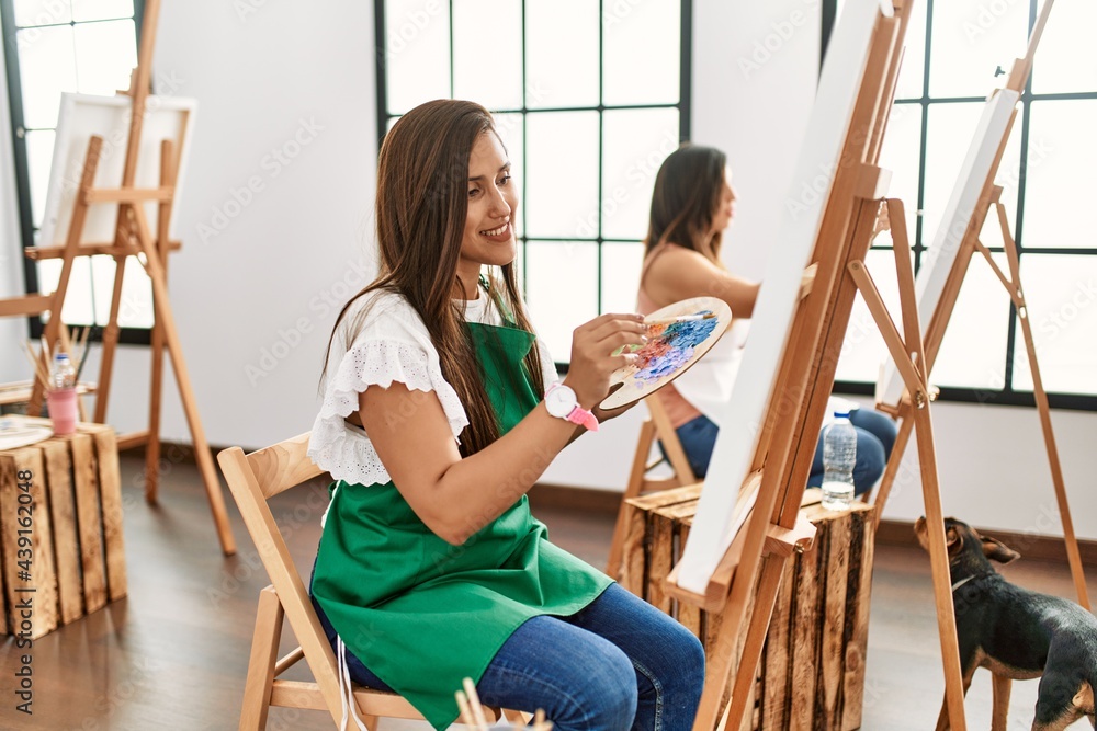 Young hispanic artist women painting on canvas at art studio smiling with happy face winking at the camera doing victory sign with fingers. number two. Young hispanic artist women painting on canvas at art studio smiling with happy face winking at the camera doing victory sign with fingers. number two.