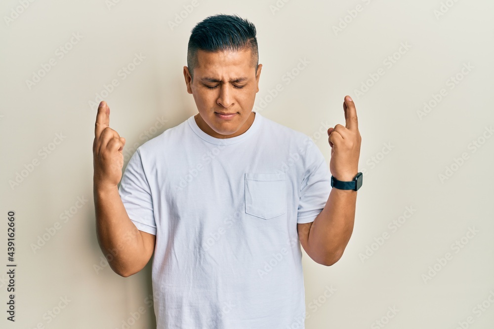 Fototapeta premium Young latin man wearing casual white t shirt gesturing finger crossed smiling with hope and eyes closed. luck and superstitious concept.