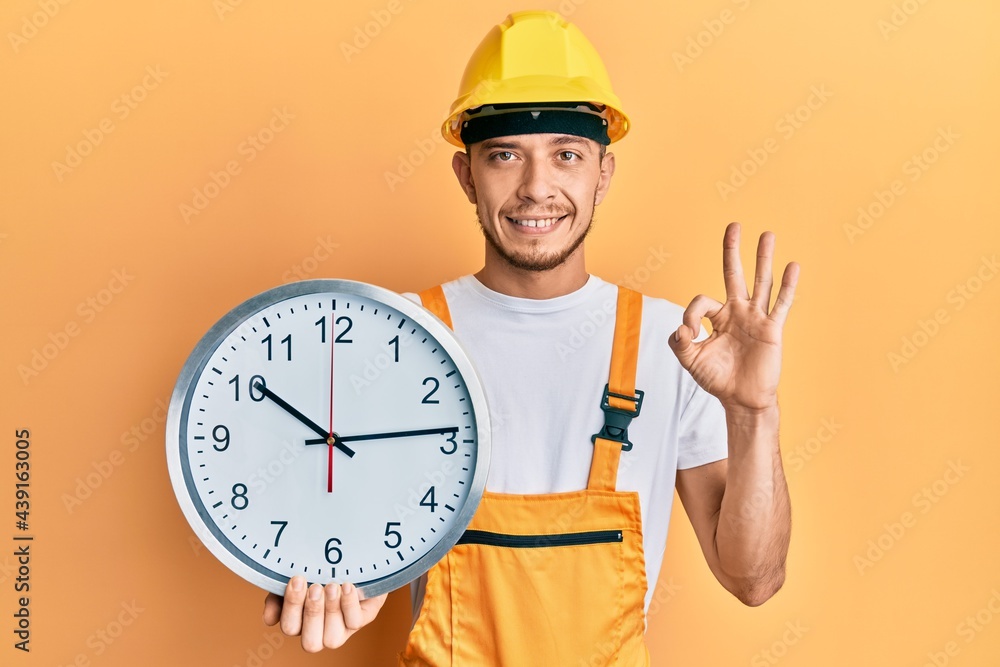 Hispanic young man wearing builder uniform and hardhat holding clock doing ok sign with fingers, smiling friendly gesturing excellent symbol Hispanic young man wearing builder uniform and hardhat holding clock doing ok sign with fingers, smiling friendly gesturing excellent symbol