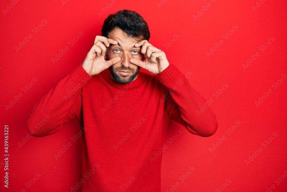 Handsome man with beard wearing casual red sweater trying to open eyes with fingers, sleepy and tired for morning fatigue Handsome man with beard wearing casual red sweater trying to open eyes with fingers, sleepy and tired for morning fatigue