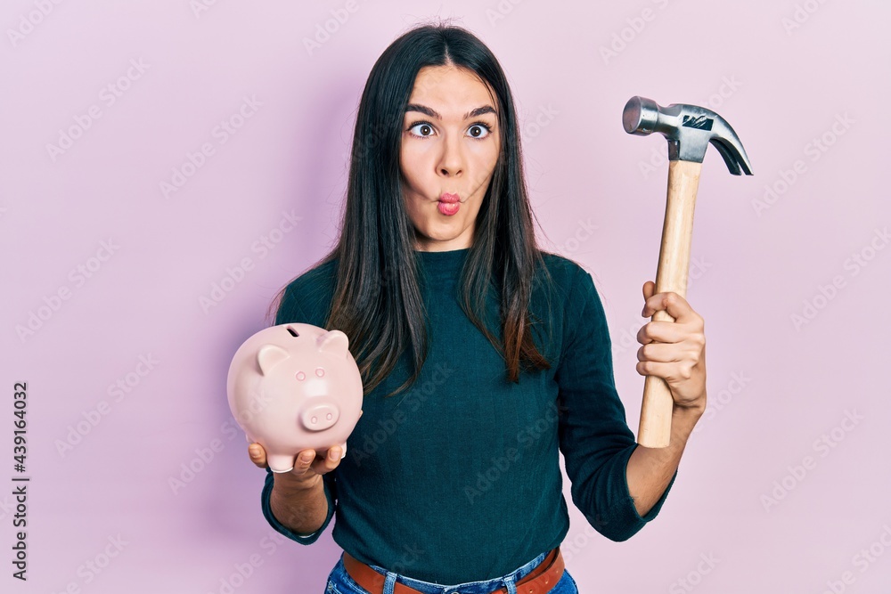 Young brunette woman holding piggy bank and hammer making fish face with mouth and squinting eyes, crazy and comical. Young brunette woman holding piggy bank and hammer making fish face with mouth and squinting eyes, crazy and comical.