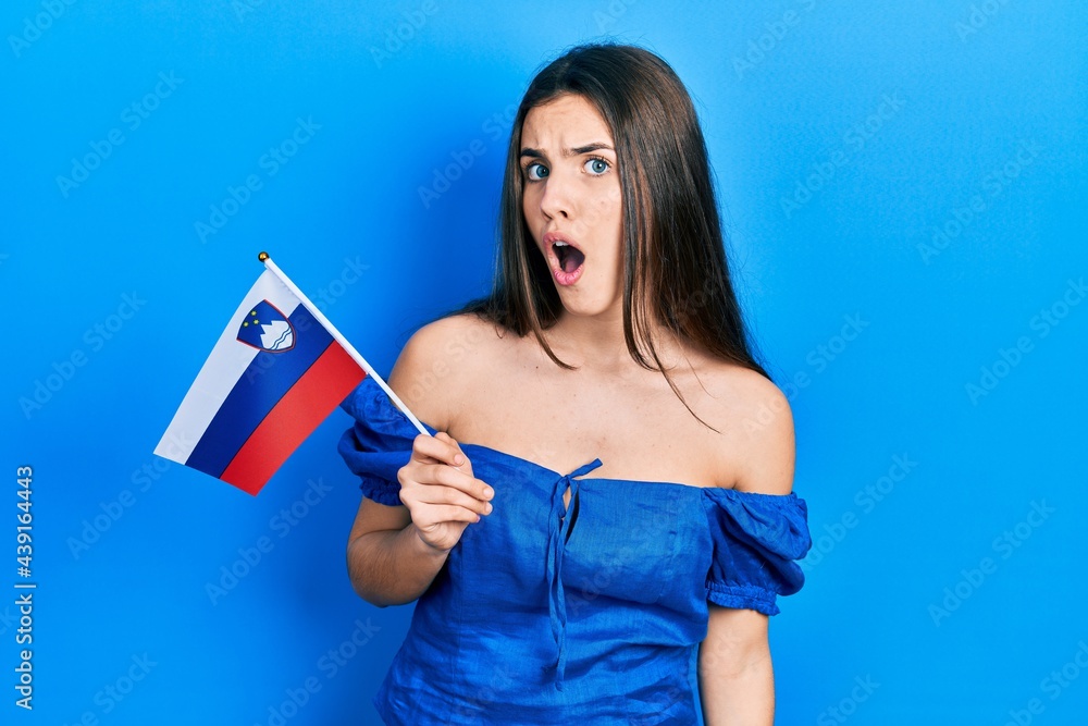 Young brunette teenager holding slovenia flag scared and amazed with open mouth for surprise, disbelief face Young brunette teenager holding slovenia flag scared and amazed with open mouth for surprise, disbelief face