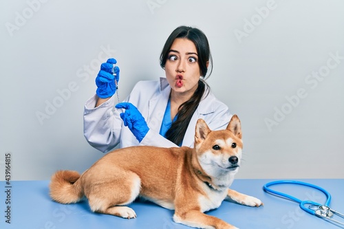 Beautiful hispanic veterinarian woman putting vaccine to puppy dog making fish face with mouth and squinting eyes, crazy and comical.