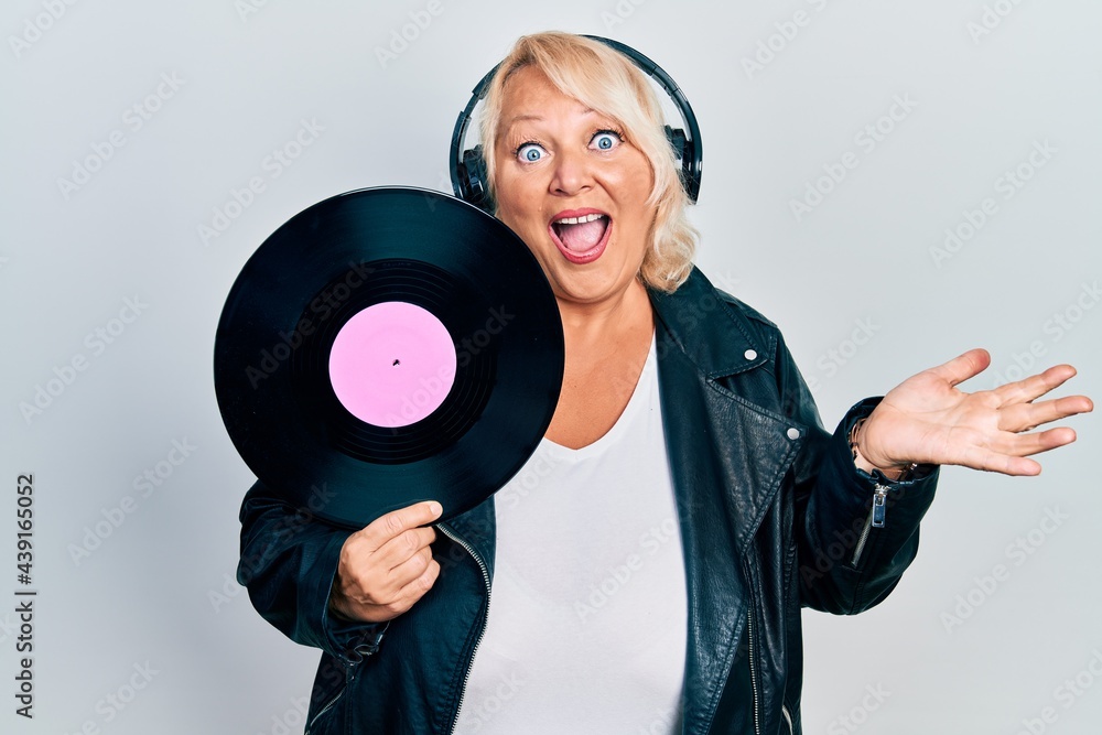 Middle age blonde woman holding vinyl disc using headphones celebrating achievement with happy smile and winner expression with raised hand Middle age blonde woman holding vinyl disc using headphones celebrating achievement with happy smile and winner expression with raised hand