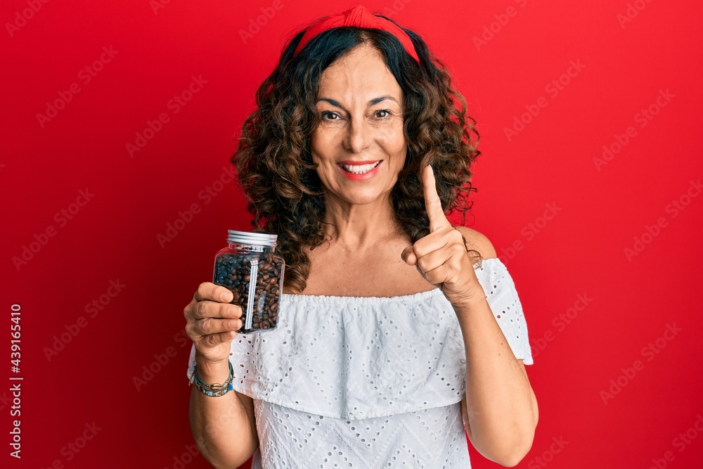 Middle age hispanic woman holding jar with coffee beans smiling with an idea or question pointing finger with happy face, number one Middle age hispanic woman holding jar with coffee beans smiling with an idea or question pointing finger with happy face, number one