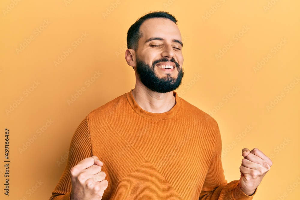 Young man with beard wearing casual winter sweater excited for success with arms raised and eyes closed celebrating victory smiling. winner concept. Young man with beard wearing casual winter sweater excited for success with arms raised and eyes closed celebrating victory smiling. winner concept.