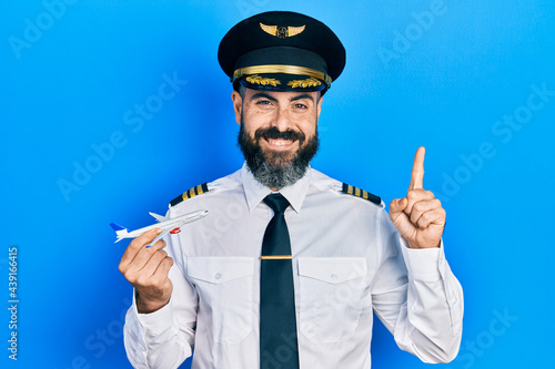 Young hispanic man wearing pilot uniform holding plane toy smiling with an idea or question pointing finger with happy face, number one photo