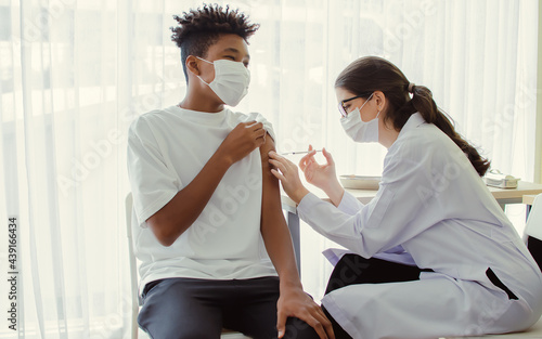 African teen man wearing face mask, getting vaccination to protect or prevent virus, getting scary vaccine injection while female caucasian doctor preparing syringe to vaccinate patient at hospital.