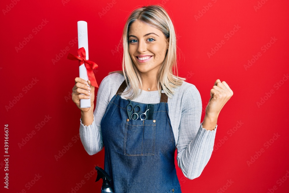 Beautiful blonde woman wearing hairdresser apron holding degree screaming proud, celebrating victory and success very excited with raised arm Beautiful blonde woman wearing hairdresser apron holding degree screaming proud, celebrating victory and success very excited with raised arm