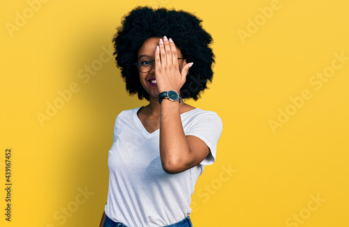 Young african american woman wearing casual white t shirt covering one eye with hand, confident smile on face and surprise emotion. © Krakenimages.com