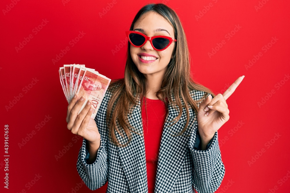 Obraz premium Young brunette woman holding 20 israel shekels banknotes smiling happy pointing with hand and finger to the side