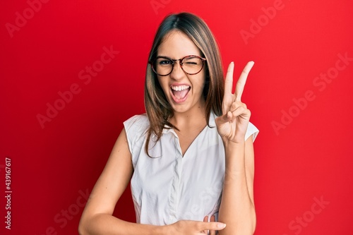 Obraz na plátně Young brunette woman wearing casual clothes and glasses smiling with happy face winking at the camera doing victory sign