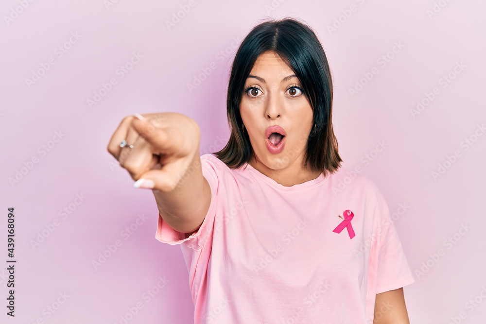 Young hispanic woman wearing pink cancer ribbon on t shirt pointing with finger surprised ahead, open mouth amazed expression, something on the front Young hispanic woman wearing pink cancer ribbon on t shirt pointing with finger surprised ahead, open mouth amazed expression, something on the front