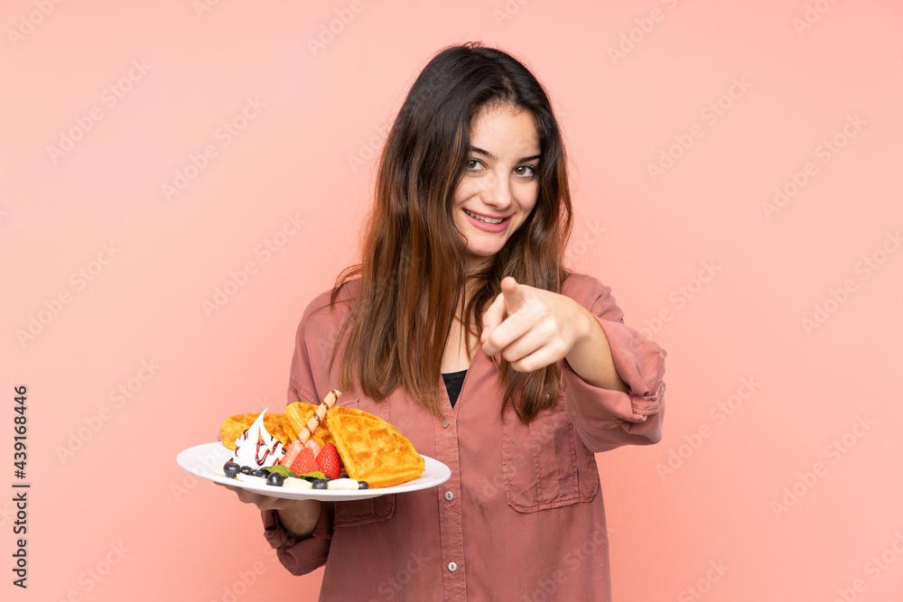 Young caucasian woman holding waffles isolated on pink background points finger at you with a confident expression