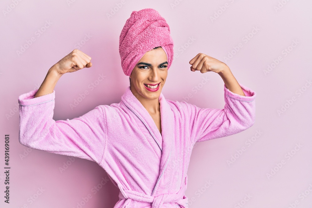 Young man wearing woman make up wearing shower towel on head and bathrobe showing arms muscles smiling proud. fitness concept. Young man wearing woman make up wearing shower towel on head and bathrobe showing arms muscles smiling proud. fitness concept.