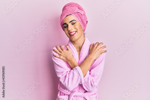 Bild auf Leinwand Young man wearing woman make up wearing shower towel on head and bathrobe hugging oneself happy and positive, smiling confident