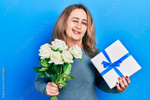 Middle age caucasian woman holding anniversary present and bouquet of flowers winking looking at the camera with sexy expression, cheerful and happy face.