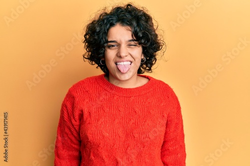 Young hispanic woman with curly hair wearing casual winter sweater sticking tongue out happy with funny expression. emotion concept. © Krakenimages.com