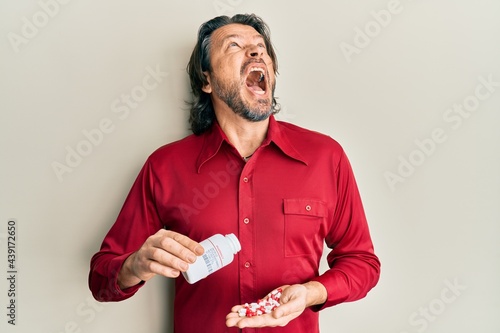 Middle age handsome man taking pills angry and mad screaming frustrated and furious, shouting with anger looking up. © Krakenimages.com